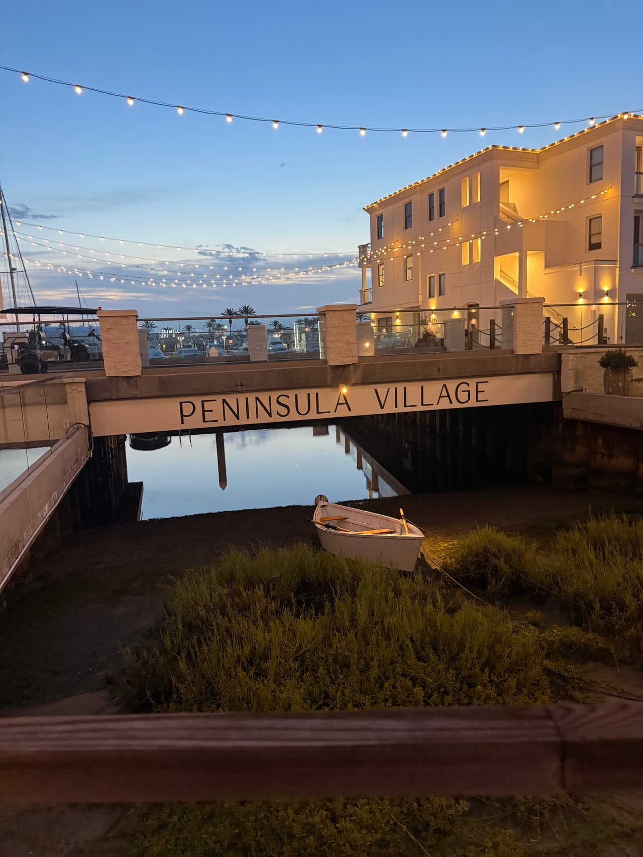 Peninsula Village sign and waterfront residences beneath string lights at dusk