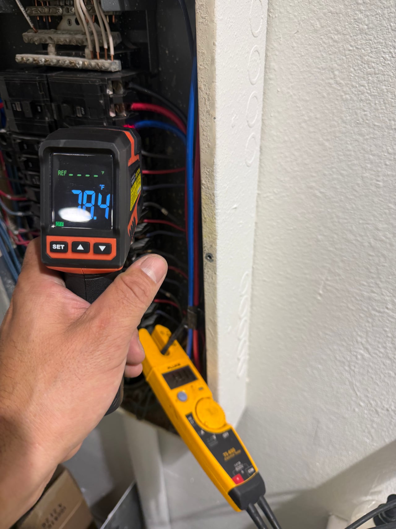 A handheld infrared thermometer and clamp meter used at an electrical panel